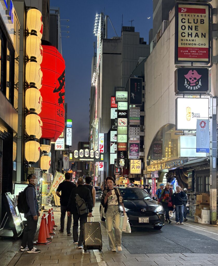 Locals walking at night in Osaka past glowing izakaya lanterns, capturing the cozy and authentic street atmosphere.