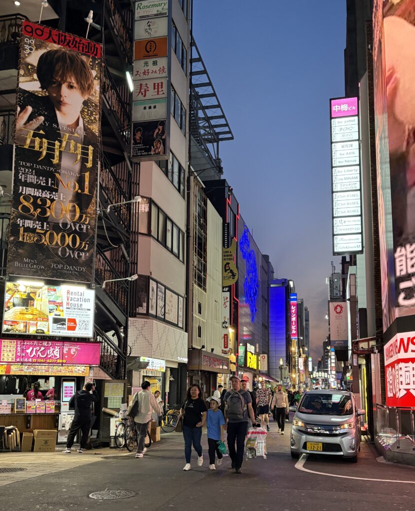 Locals walking through a bustling Osaka street at night, surrounded by illuminated signs and colorful advertisements.