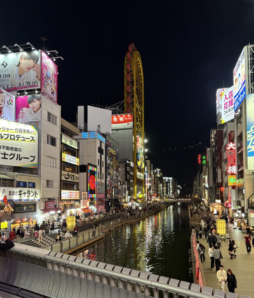 Nighttime river view in Osaka with bright neon signs and crowds of tourists walking along the waterfront.
