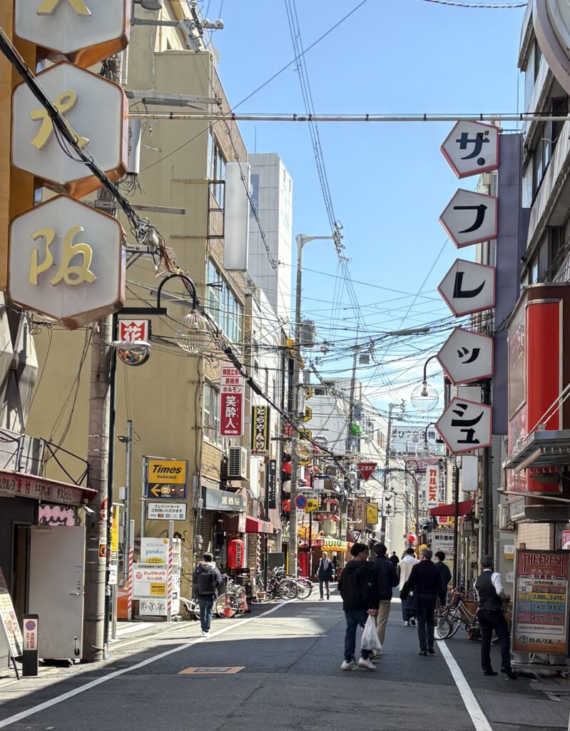 Sunny daytime view of a quiet street in Osaka lined with local storefronts and signage