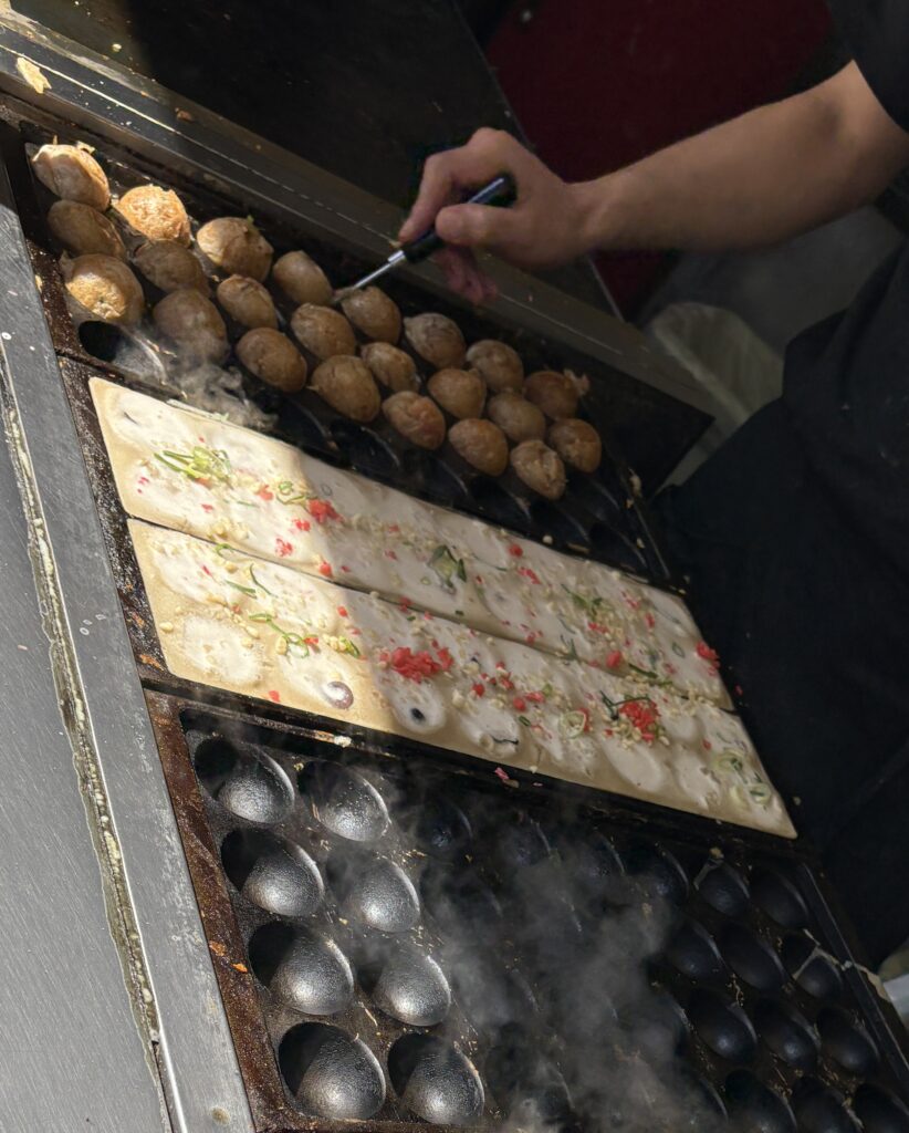 Golden takoyaki octopus balls sizzling on a cast iron grill at a busy Osaka street food stall.