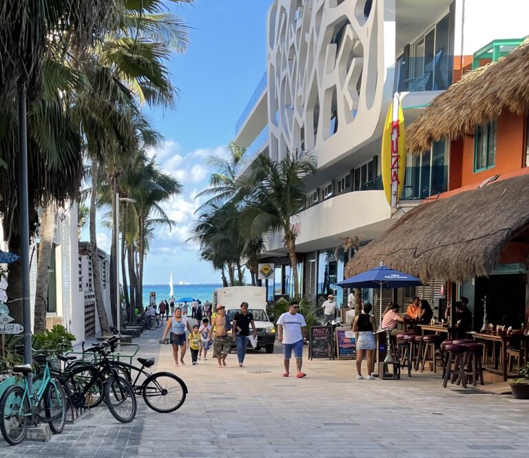 People walking down a vibrant street in Playa del Carmen’s Centro area with bikes parked and the sea visible in the distance, restaurants lining the sides.