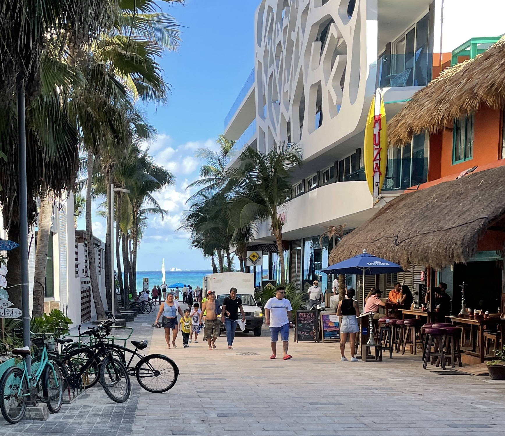 People walking down a vibrant street in Playa del Carmen’s Centro area with bikes parked and the sea visible in the distance, restaurants lining the sides.