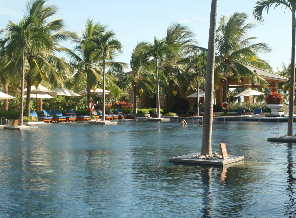 Large resort swimming pool at An Bang Beach in Hoi An, Vietnam, surrounded by palm trees and lounge chairs under a clear blue sky.