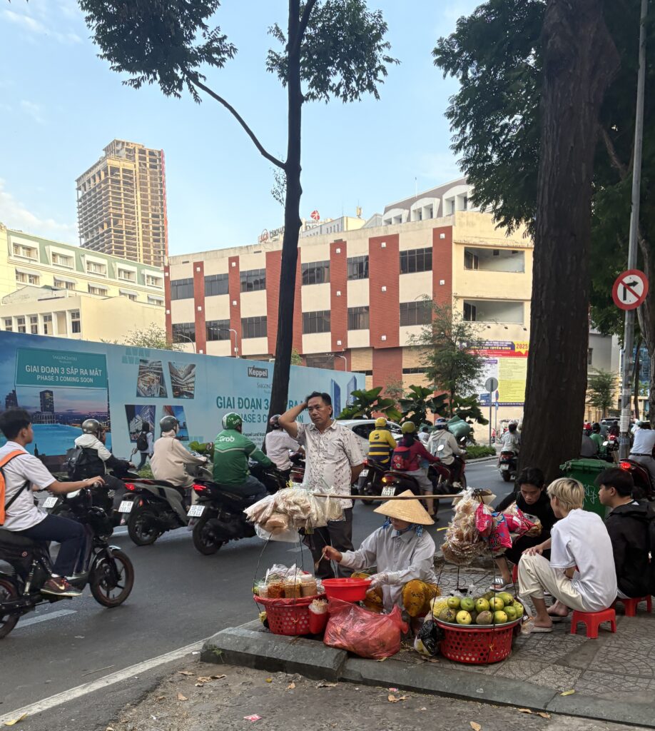 Local street vendor sitting on a small stool serving food to customers with scooters passing by in Ho Chi Minh City