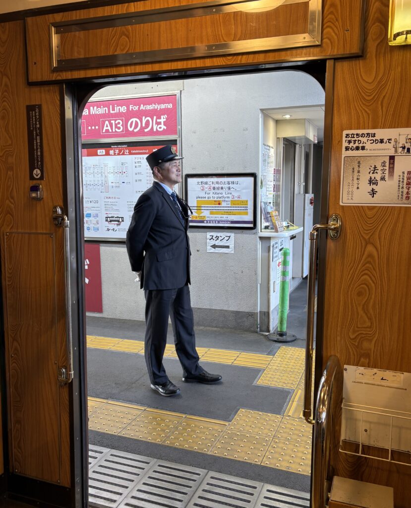 Train conductor in classic uniform operating a traditional Kyoto streetcar, seen from inside with door open.