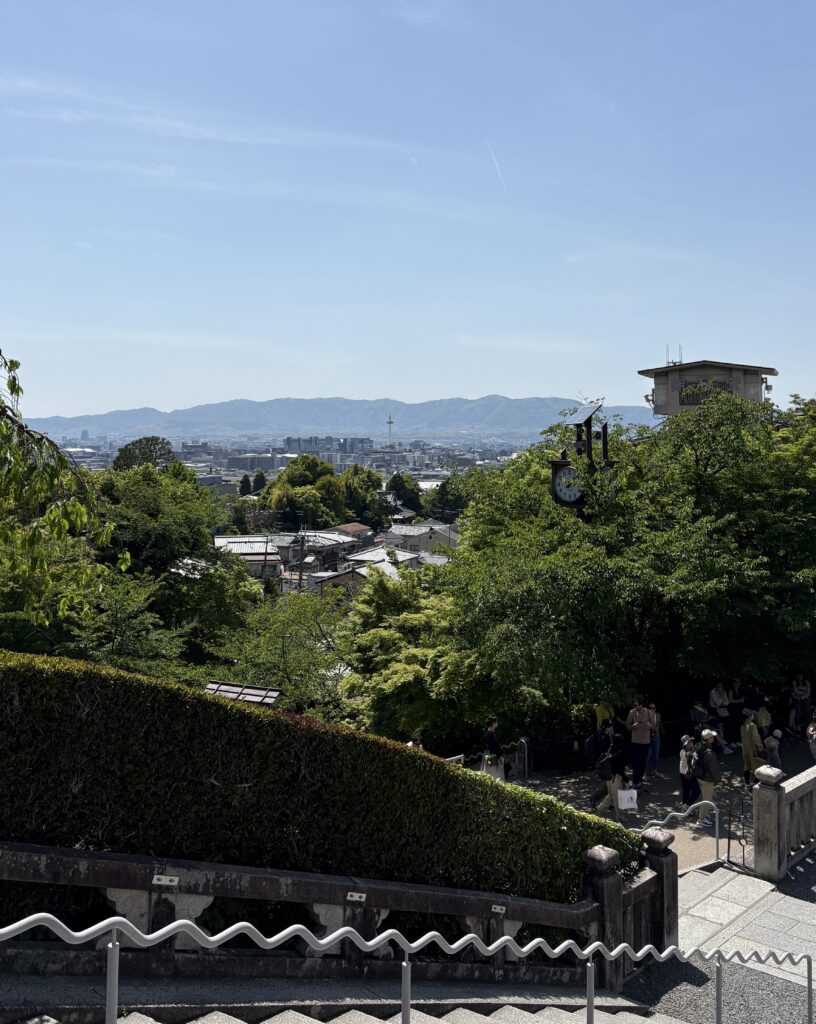 Tourists walking along a stepped path in Kyoto with panoramic views of the city and distant mountains.