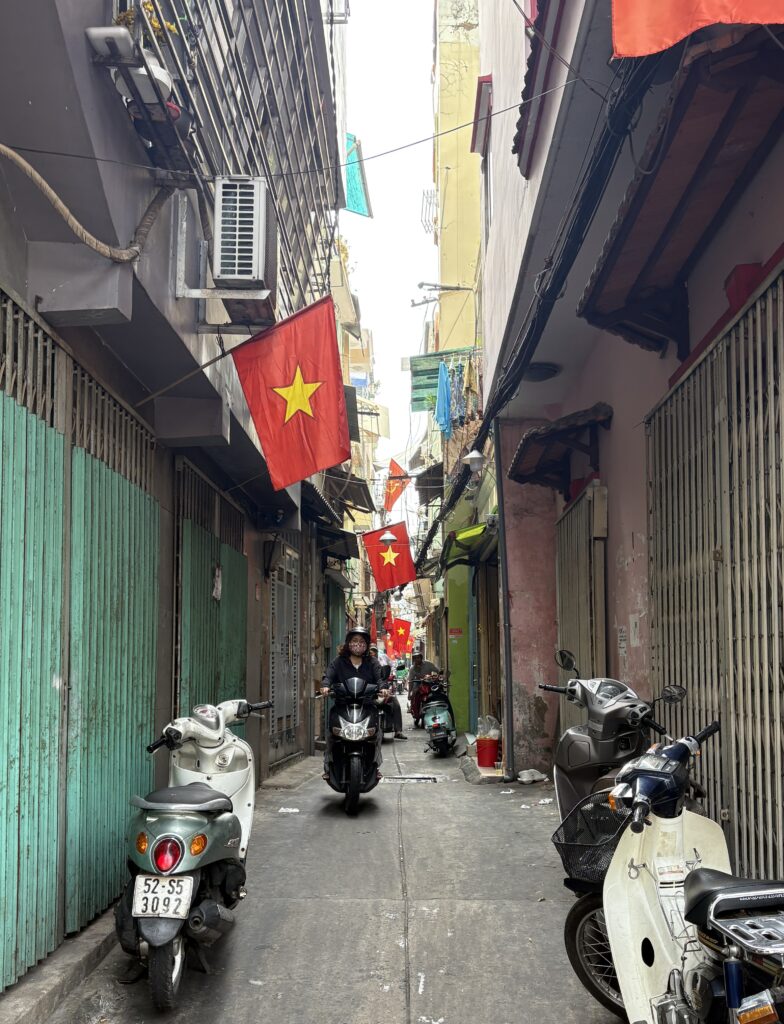 A narrow alleyway in Ho Chi Minh City decorated with Vietnam flags, with scooters parked on the side and a woman riding by on a motorbike.