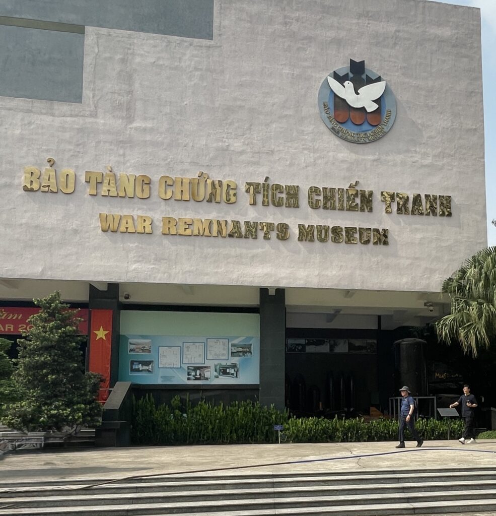 Front entrance of the War Remnants Museum in Ho Chi Minh City with visitors outside and Vietnamese flags displayed.