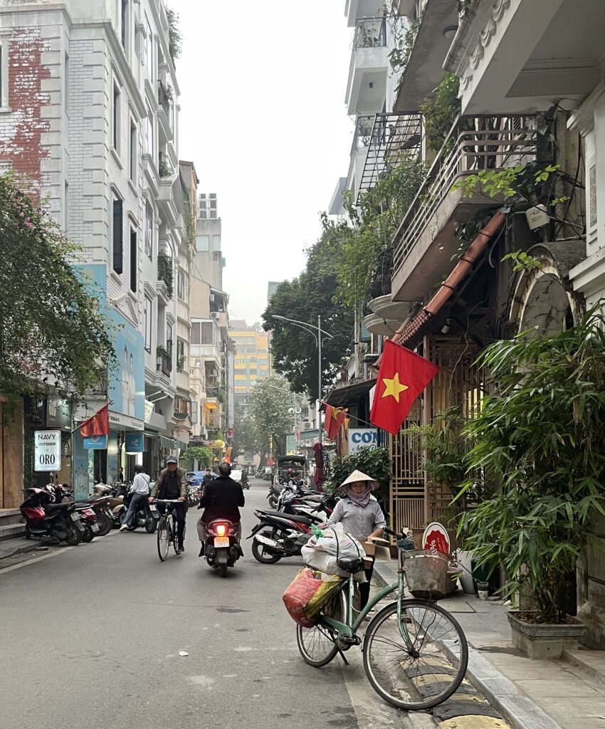 Featured image for where to stay in Hanoi: A woman selling fruit from her bicycle with the Vietnamese flag nearby, captured on a quiet Hanoi street.