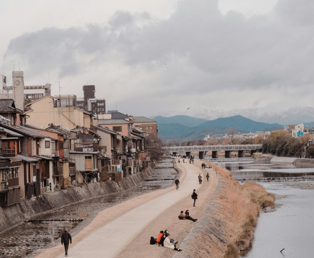Locals sitting by Kyoto’s Kamo River during winter, with bare trees and mountain scenery in the background.