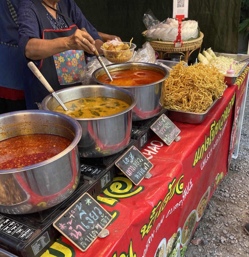 A woman at the Chiang Mai Sunday Market preparing a fresh bowl of Khao Soi at her street food stall for a customer.