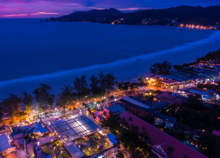 Aerial view of Patong Beach at night with bright street lights, sandy shoreline, and mountains in the background in Phuket, Thailand.