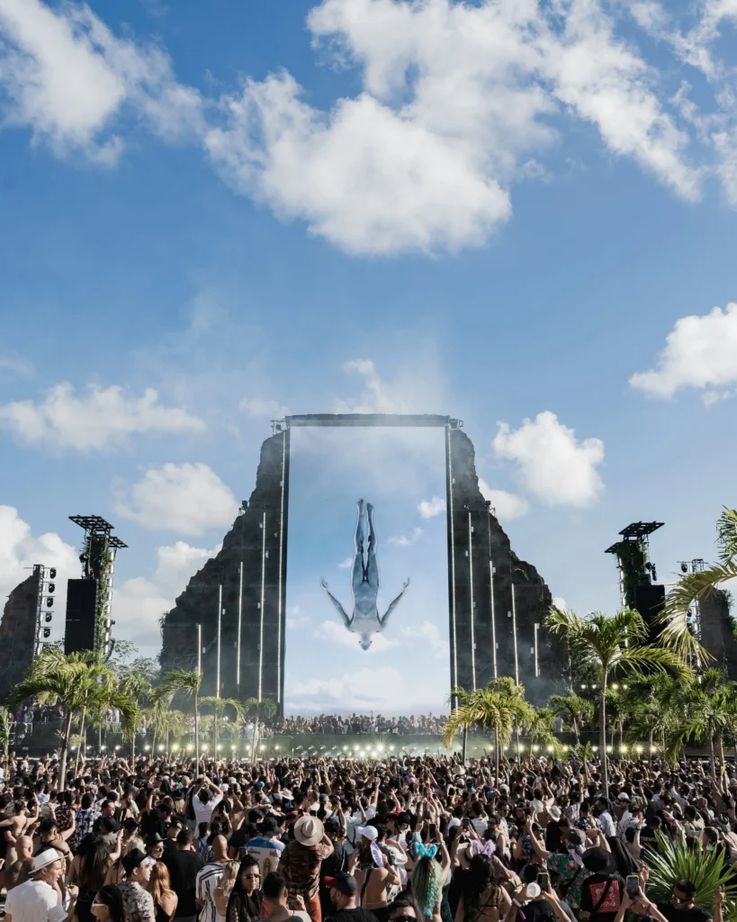 Daytime crowd at Afterlife Tulum festival with large digital display under a sunny sky
