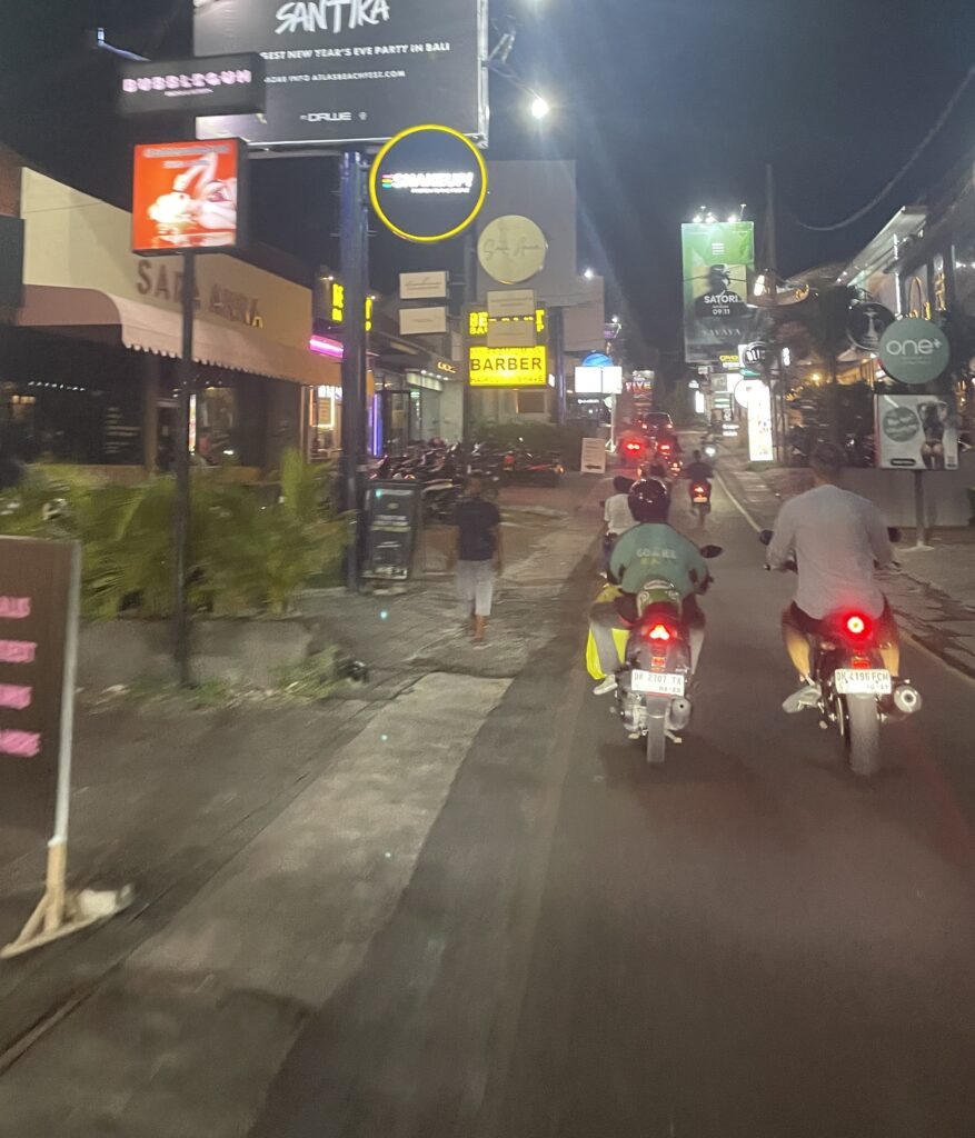 Poor infrastructure in Bali at night with scooters passing by, narrow sidewalks, limited walking space, and visible potholes