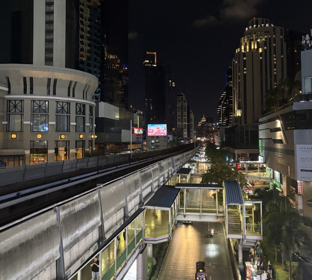 Elevated night view of Bangkok city from a BTS station, showing the skytrain railway and illuminated skyline.