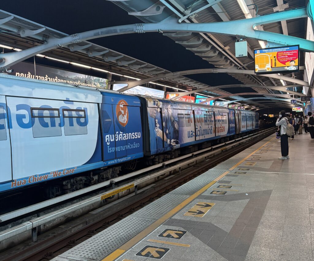 Close‑up of a BTS Skytrain in Bangkok at night with locals and tourists waiting on the platform.