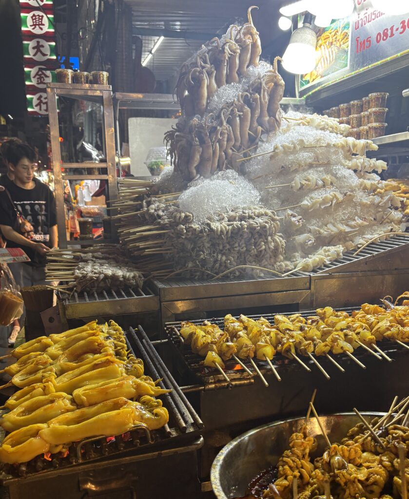 Piles of raw squid stacked beside an electric grill cooking multiple squid skewers at a busy street food stall in Bangkok’s Chinatown.