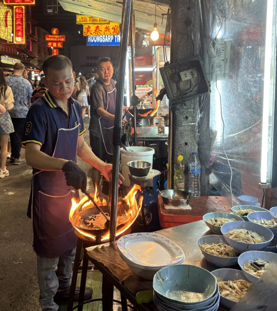 A street food vendor cooking with a fiery wok in Bangkok’s Chinatown, with plates of food being prepared on the side.