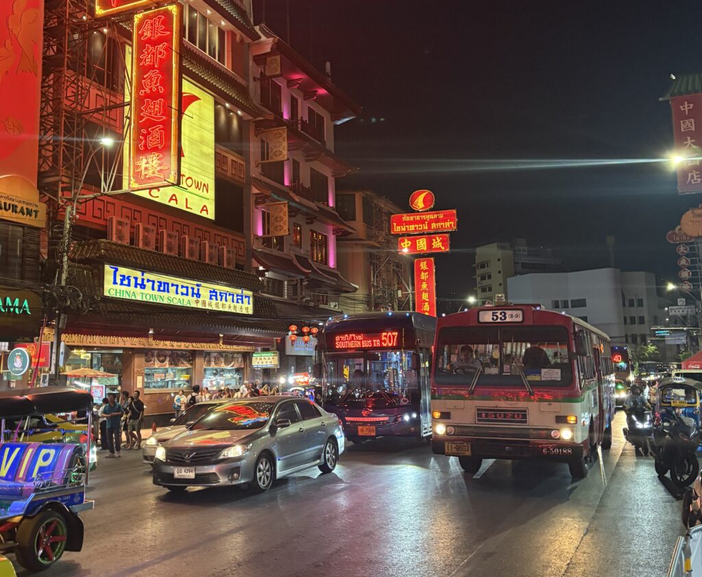 A Bangkok public bus passes through Chinatown at night, with glowing neon signs, bustling streets, and locals and tourists walking nearby.