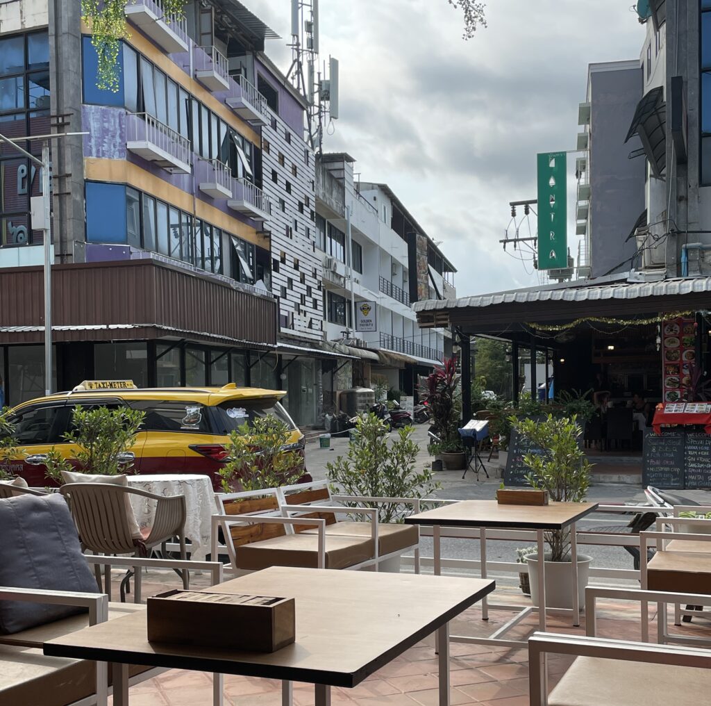 Restaurant tables along a street in Chaweng, Koh Samui, with a taxi passing by on a cloudy midday
