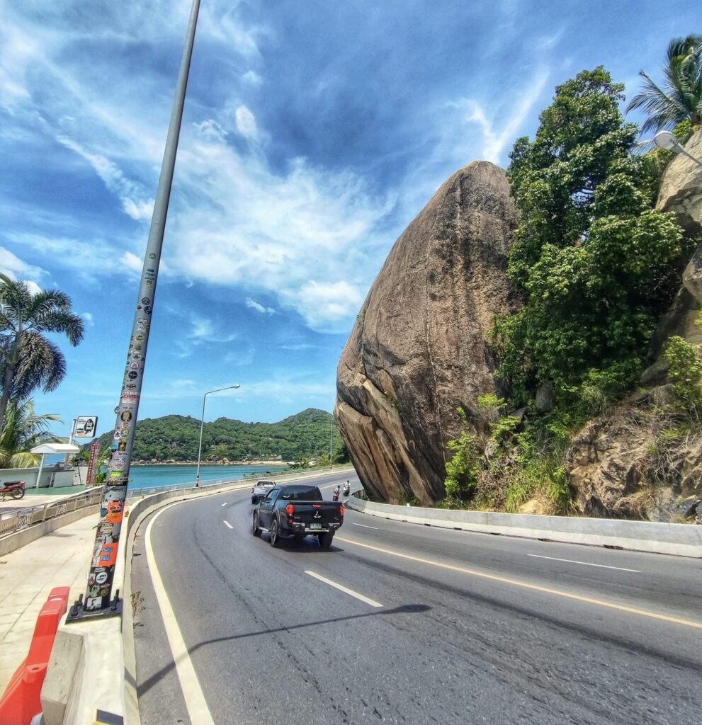 Scenic coastal road in Koh Samui with the sea on the left and a large rock on the right on a clear day