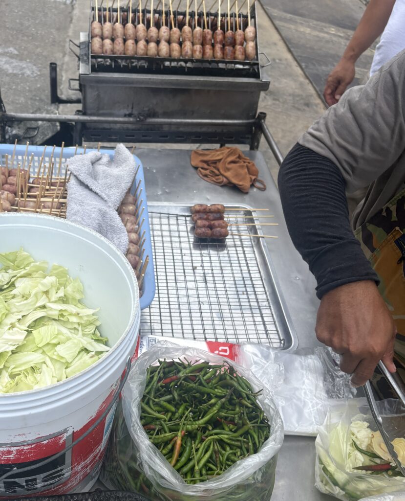 A Thai street food vendor grilling moo ping skewers with chilies visible on the counter, showcasing authentic Bangkok night market vibes.