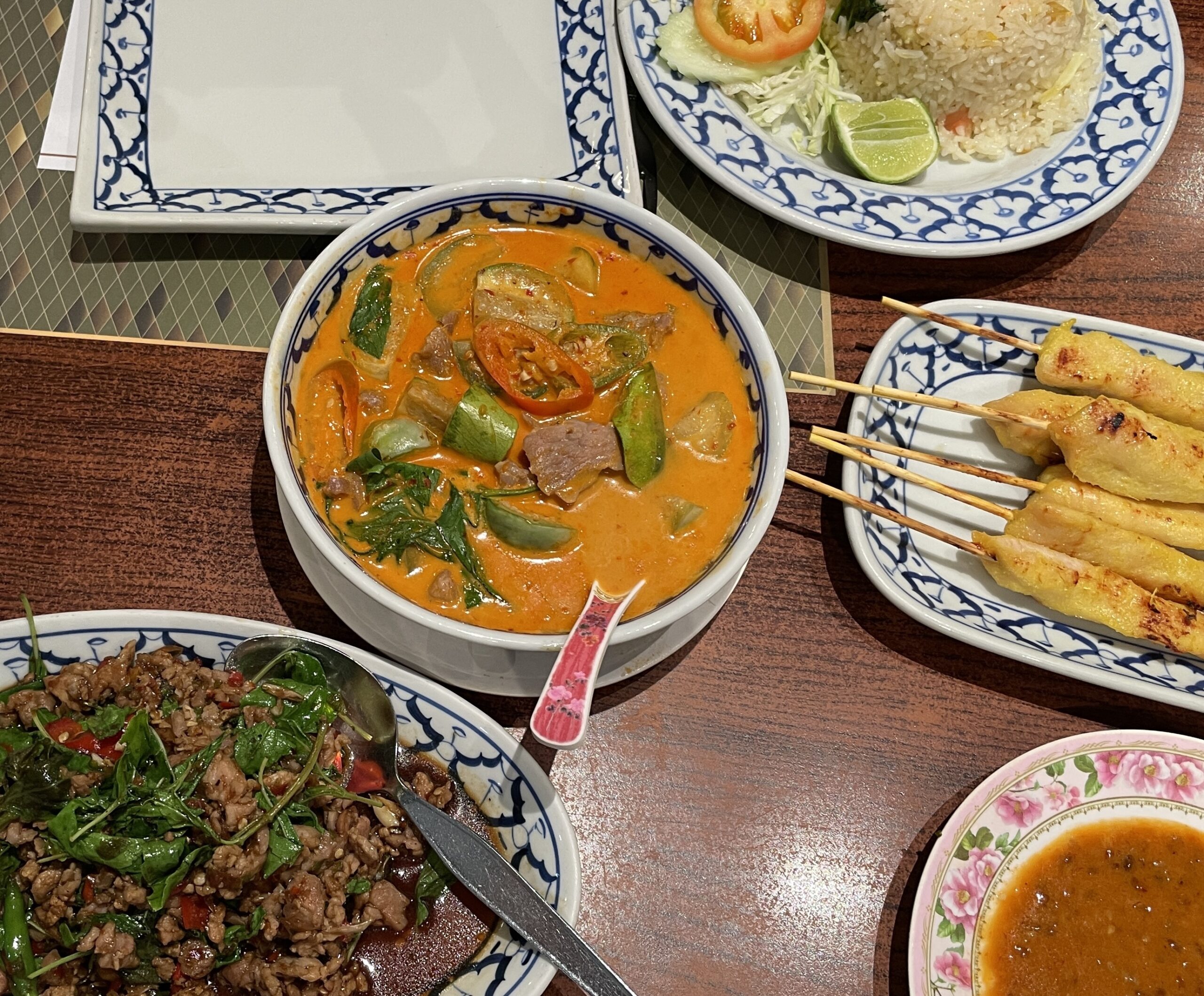 A table of classic Thai dishes including Pad Kra Pao with fried egg, chicken satay with peanut sauce, and creamy Panang curry.