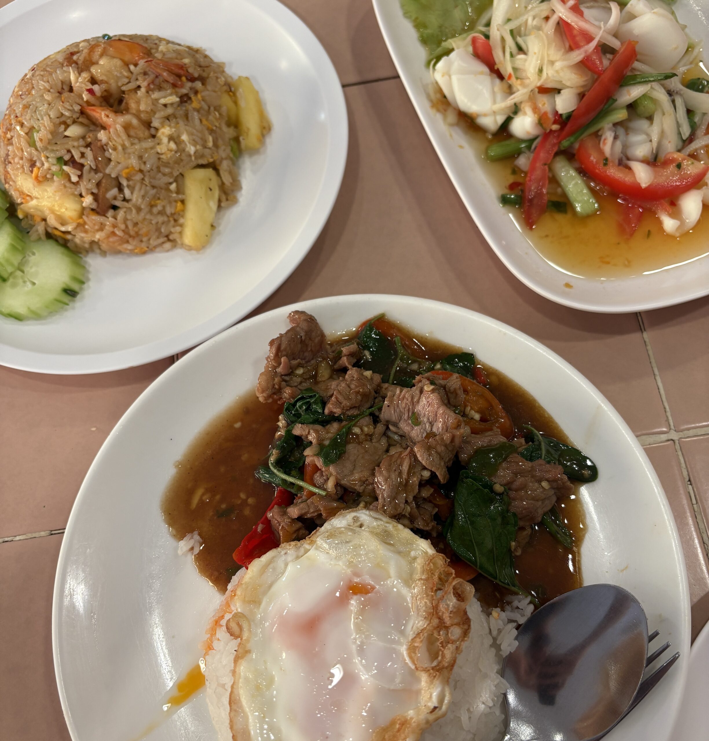 A table spread featuring Pad Kra Pao, Thai squid salad, and Tom Yum fried rice, showcasing vibrant Thai flavors.