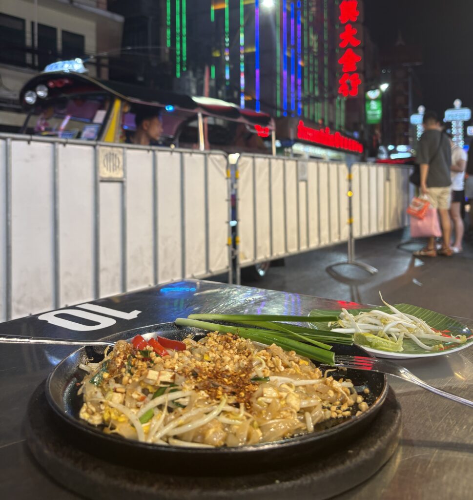 Close-up of Pad Thai on a plate with a tuk-tuk and tourists in Bangkok’s Chinatown
