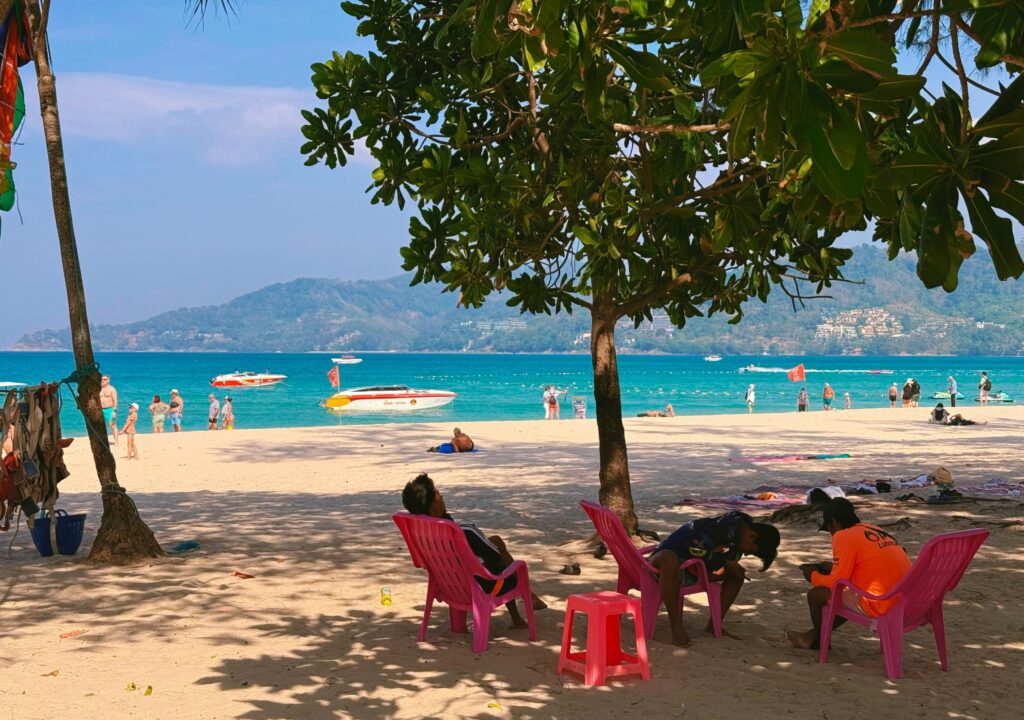 Sunny day on Patong Beach in Phuket, Thailand, with three local men offering jet ski rentals, tourists on the sand, and a boat floating in the clear blue water.
