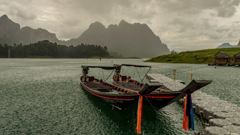 Two long-tail boats on the water in Thailand during the rainy season, with heavy rain falling and mountains in the background.