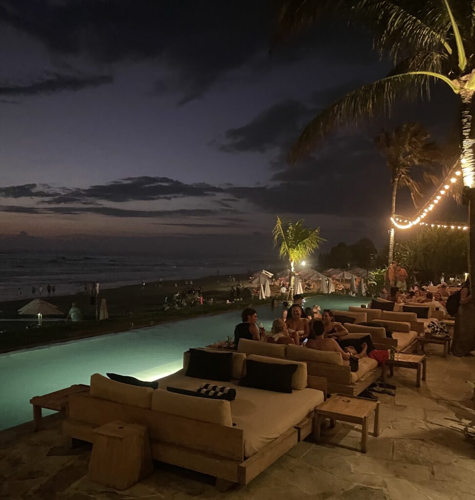 Nighttime at The Lawn beach club in Canggu with tourists dining, lounging by the pool, and the beach visible in the distance