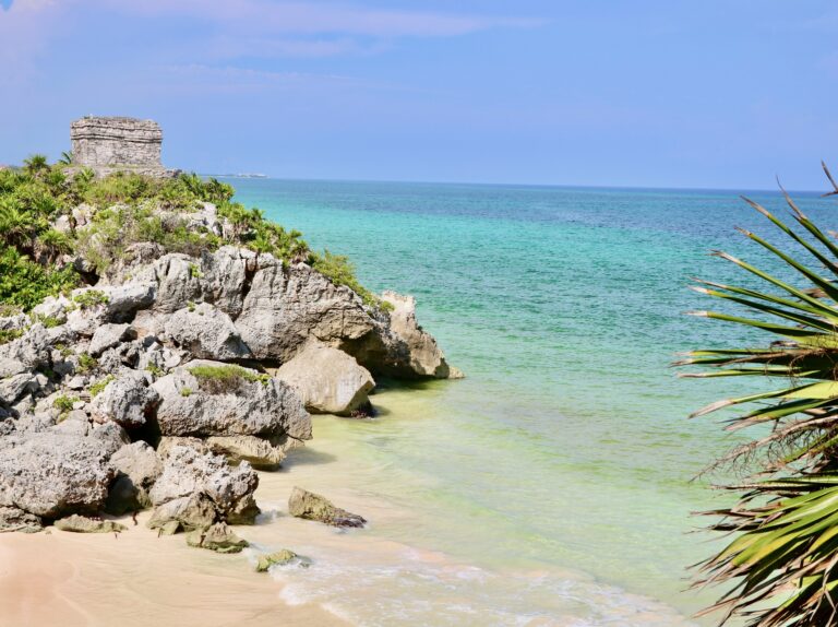 Scenic Tulum beach with calm blue water, palm trees, and an ancient sculpture along the shoreline