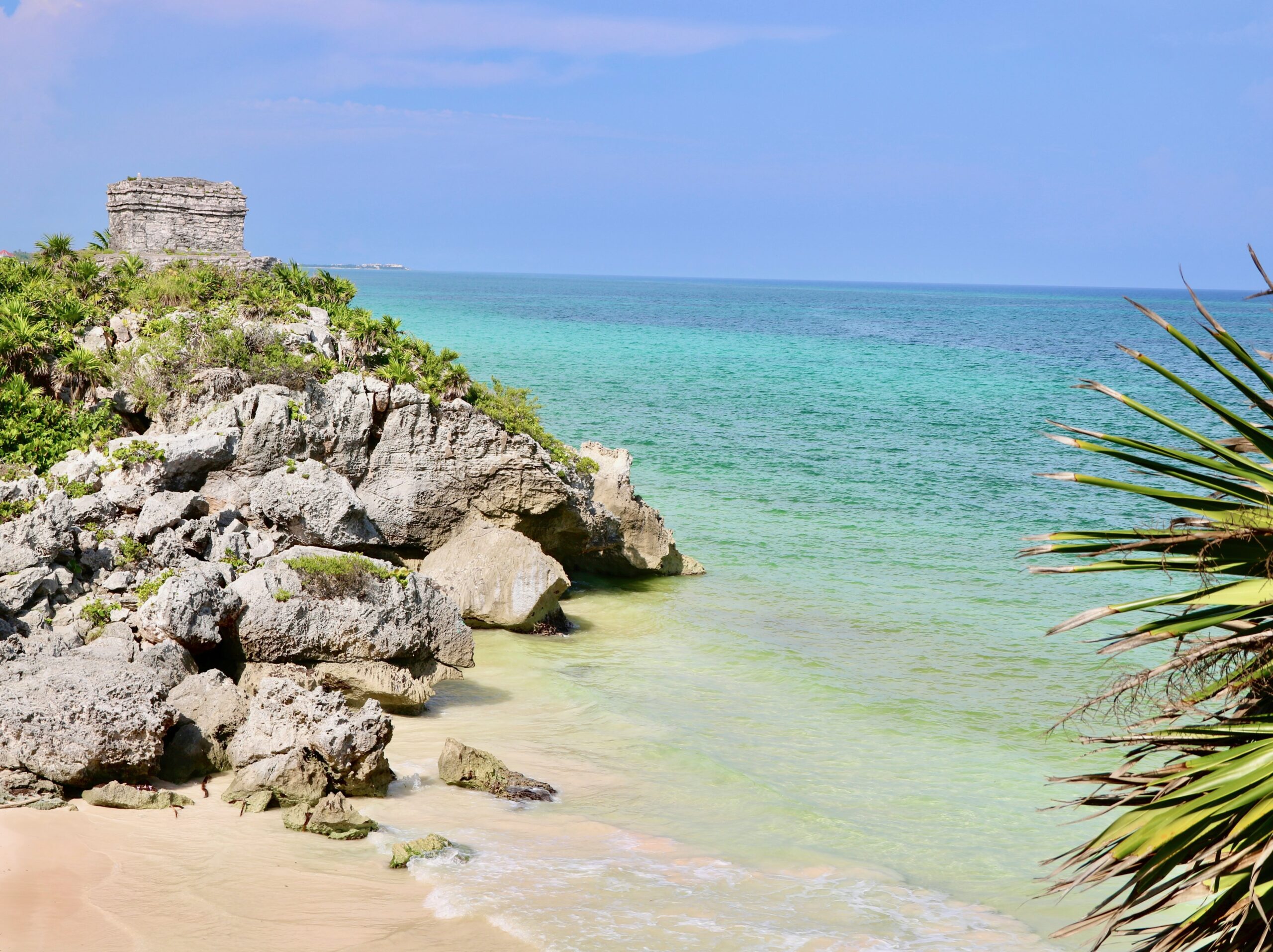 Scenic Tulum beach with calm blue water, palm trees, and an ancient sculpture along the shoreline