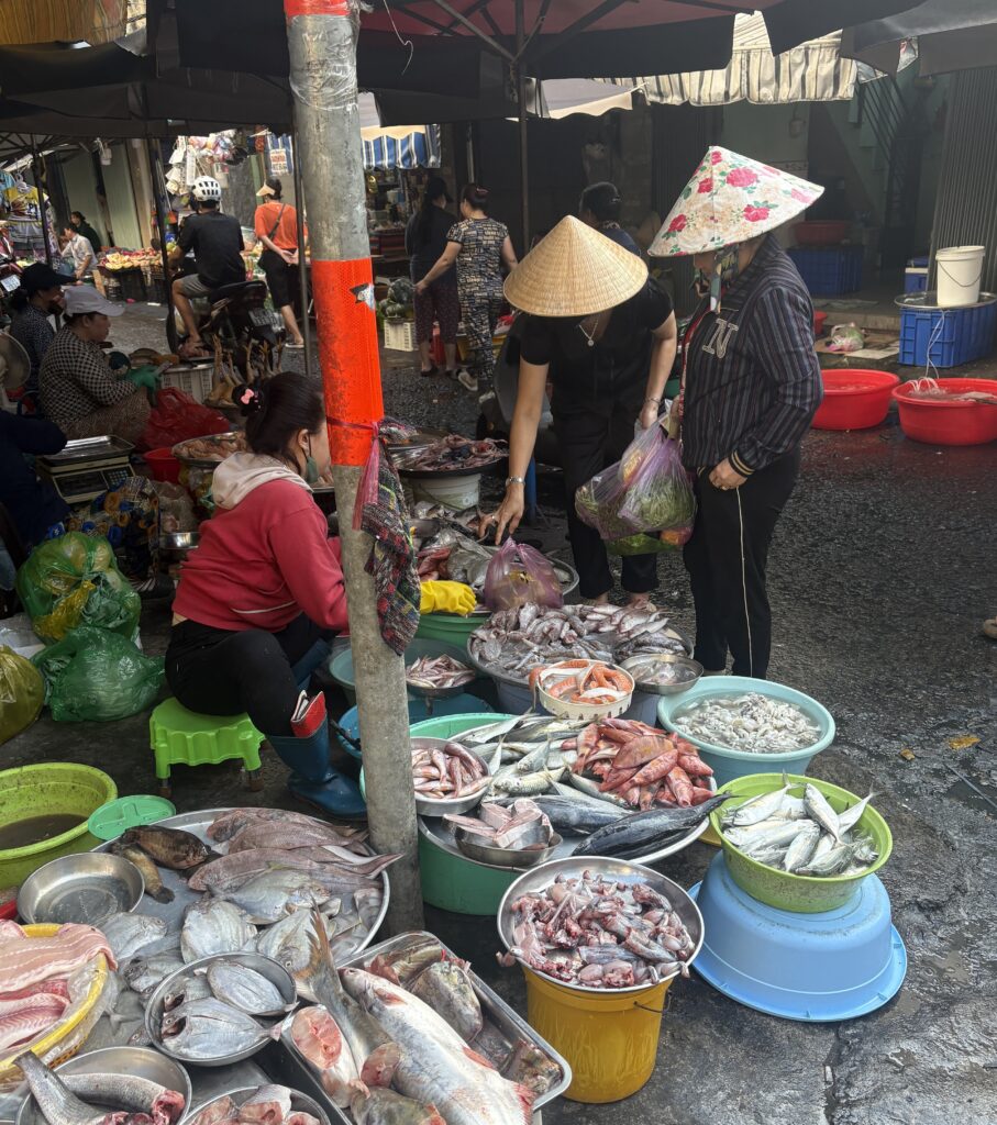 Two local women wearing traditional Vietnamese hats shopping for fresh fish at a market in Saigon.
