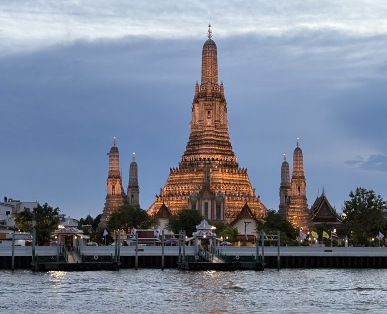 Evening sunset view of Wat Arun in Bangkok beautifully lit up, seen from the riverside with a warm glow over the Chao Phraya River.