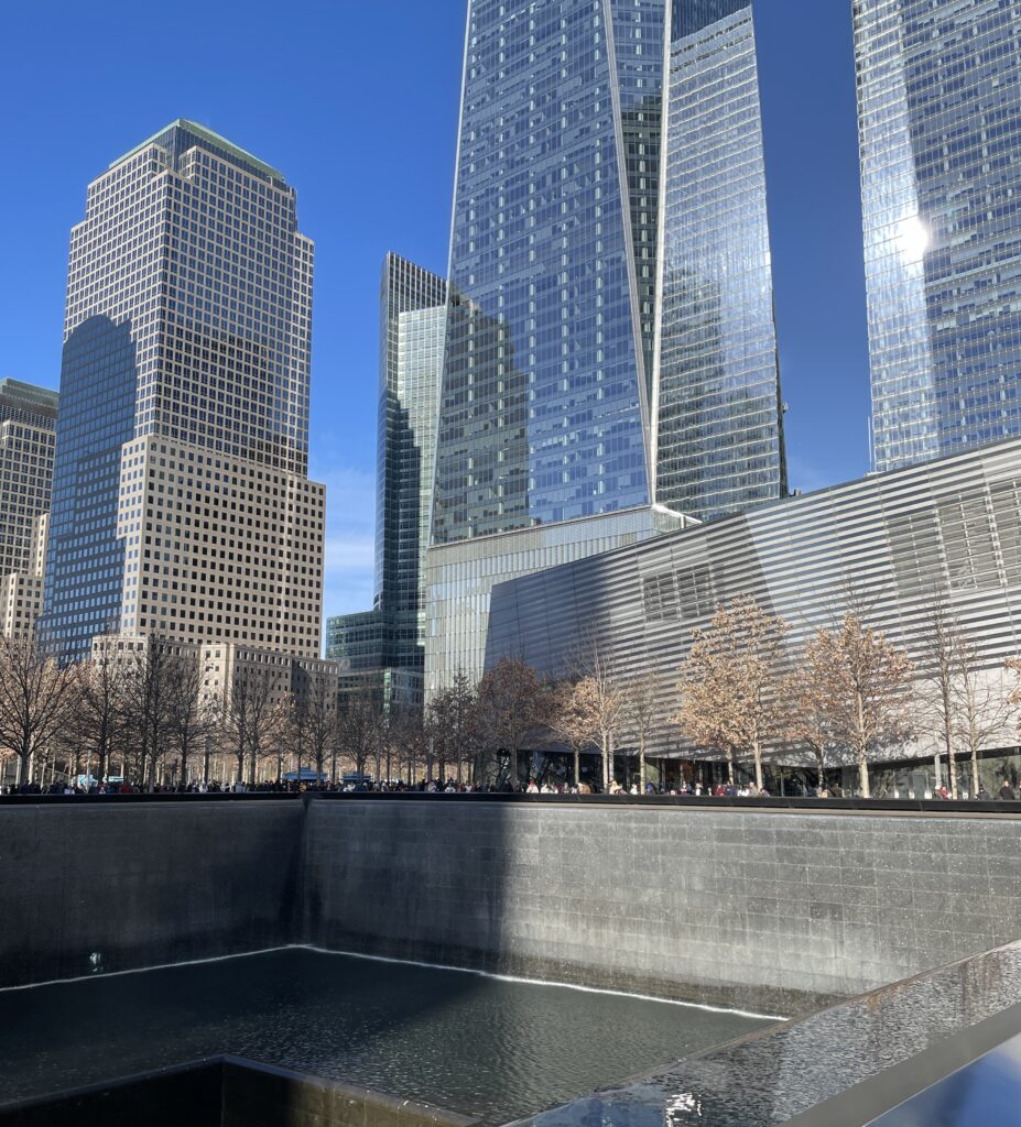 The 9/11 Memorial in New York City on a sunny day, with reflections in the water and names engraved around the fountain