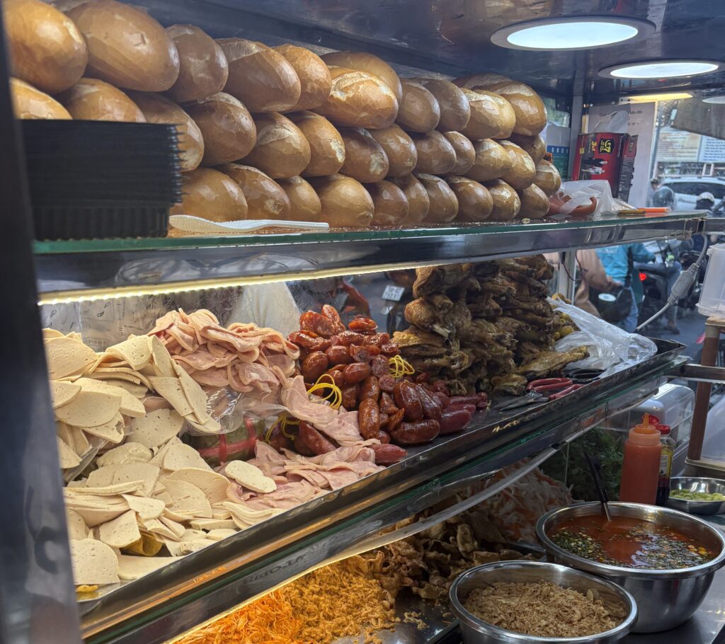 Close-up of a bánh mì stall in Saigon, showing fresh bread stacked on top and a clear display of meats, toppings, and fillings under the lights.