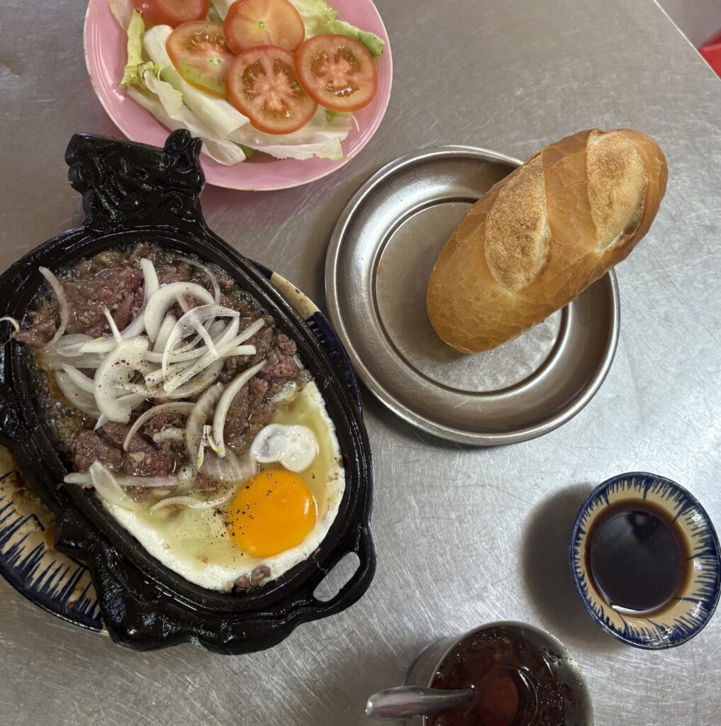 A sizzling plate of bò né with bread on a metal table in Saigon, a popular Vietnamese steak and egg dish.