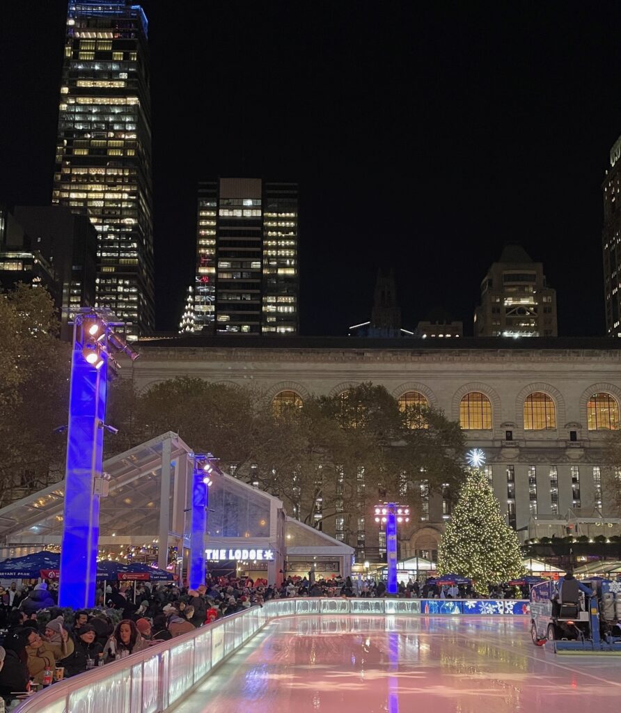 Bryant Park ice rink during Christmas with locals and tourists skating and the holiday market in the background.
