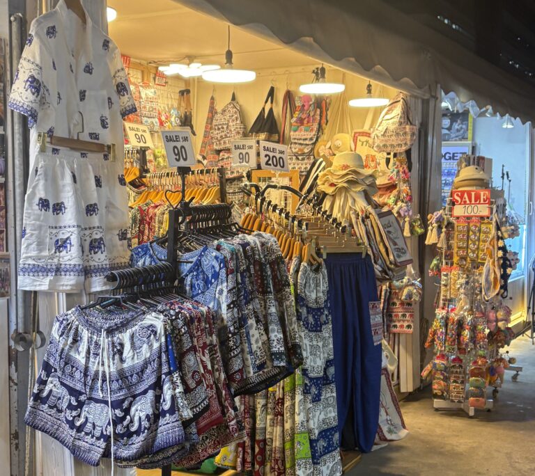 Elephant pants and shirts on display with bags in the background at Jodd Fairs night market in Bangkok.