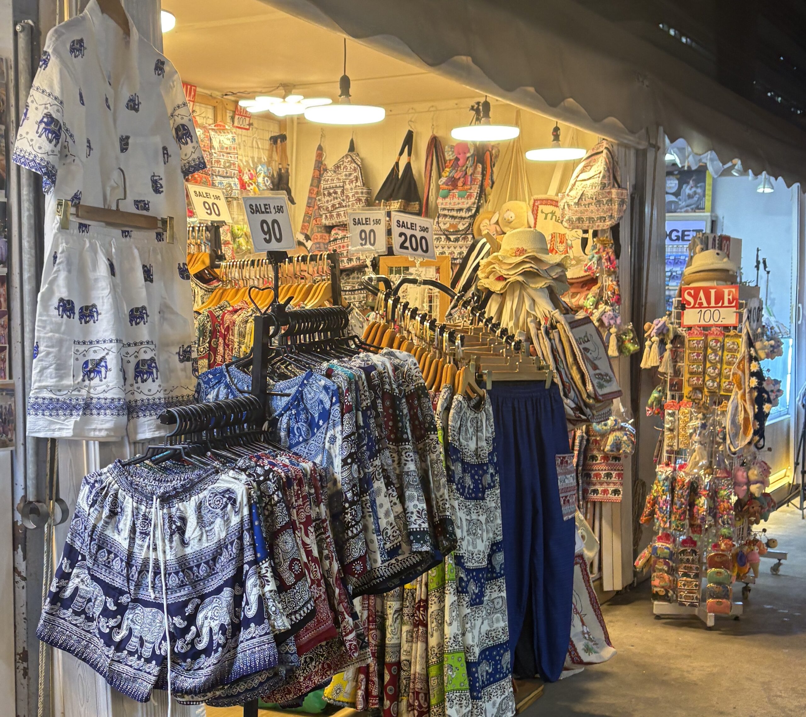 Elephant pants and shirts on display with bags in the background at Jodd Fairs night market in Bangkok.