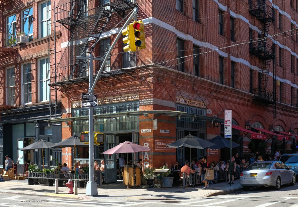 Locals and tourists dining at a lively Italian restaurant on a New York City street corner in summer.