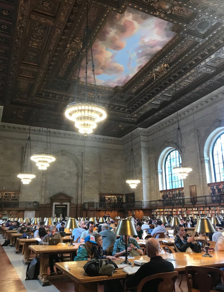 Inside the New York Public Library with people studying, reading, and browsing magazines in the grand reading room.
