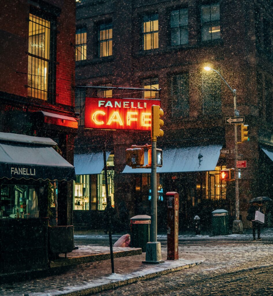 Brightly lit café sign glowing on a snowy night in New York City during January, snow covering the streets.