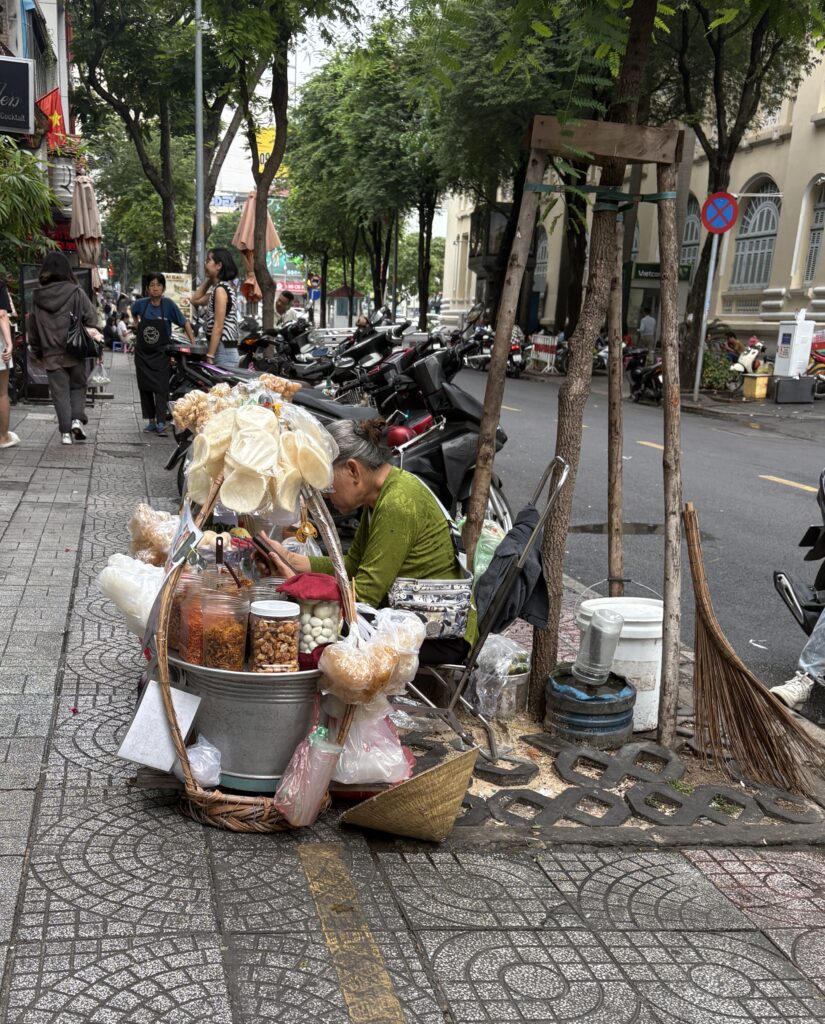 A woman vendor selling on the sidewalk in Saigon with scooters passing by and visible sidewalk damage ahead.
