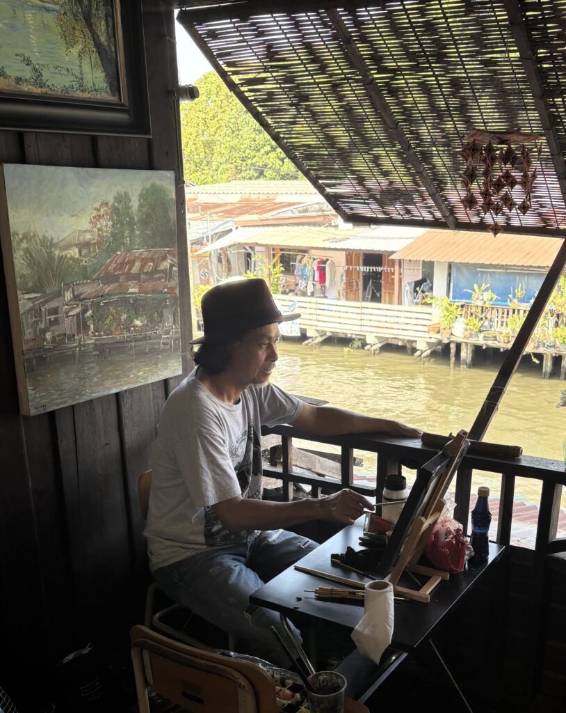 A Thai artist painting with his artwork displayed behind him at a riverside area in Thailand.