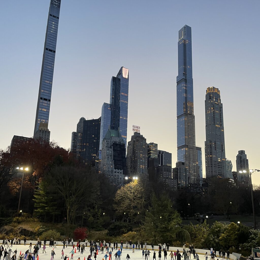 Locals and tourists skating at Wollman Rink in Central Park during early evening, with Billionaires’ Row skyscrapers in the background
