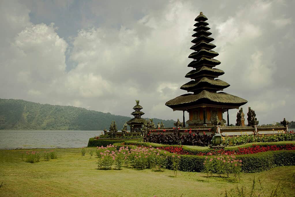 Traditional Balinese temple surrounded by lush greenery with water and distant hills in the background.